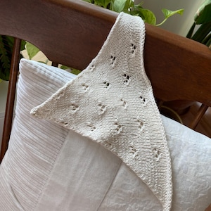 May include: A cream-coloured crocheted shawl with a delicate openwork pattern. The shawl is draped over a white cushion and a wooden chair.