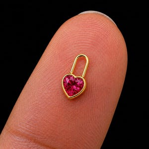 Puede incluir: Un pequeño dije de candado en tono dorado con una piedra preciosa de color rosa vibrante, tallada en forma de corazón y facetada en el centro. El marco de metal es liso y pulido, formando una clásica curva en U sobre el engaste.