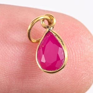 May include: A gold pendant featuring a magenta, teardrop-shaped gemstone. The gemstone is faceted and set in a gold bezel. The pendant is held by a finger. It has a loop at the top for a chain, suitable for a necklace.