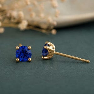 Könnte beinhalten: Ein Paar Ohrstecker mit runden, im Brillantschliff geschliffenen blauen Edelsteinen in vierzinkigen Goldfassungen. Ein Ohrring ist von vorne zu sehen, während der andere im Seitenprofil positioniert ist und den Stift sowie den Verschluss zeigt.