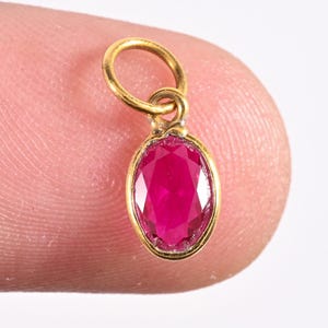 Puede incluir: Un pequeño colgante dorado con una piedra preciosa de rubí ovalada. El rubí está engastado en un bisel de oro, con un lazo para sujetarlo. La piedra preciosa es de un color rojo intenso. Una joya delicada.