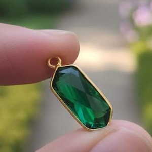 Puede incluir: Un colgante dorado con una gema verde esmeralda rectangular. El colgante tiene un corte facetado, que capta la luz. El fondo es una escena borrosa al aire libre con vegetación.