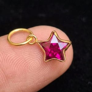 Può includere: Un ciondolo a forma di stella color oro con un centro di pietra preziosa magenta sfaccettata. Il ciondolo è sospeso da un piccolo anello circolare e viene tenuto tra due dita su uno sfondo scuro.