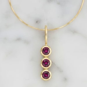 Pode incluir: Um colar com pingente de ouro com três pedras preciosas redondas, vermelho escuro, engastadas em biséis individuais. O pingente pende de uma delicada corrente de ouro, criando um design simples, mas elegante. O colar é exibido em um fundo de mármore branco.