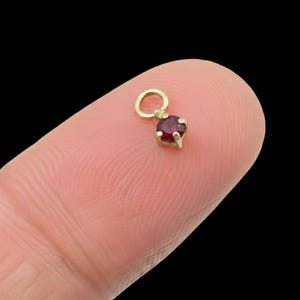 May include: A small, gold-coloured charm featuring a round, deep red gemstone. The charm has a loop at the top for attaching to a necklace or bracelet. The gemstone is set in a gold bezel.