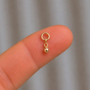 May include: A small gold-toned jewellery charm featuring a circular jump ring connected to a tiny, polished spherical bead. The charm has a smooth, reflective metallic finish and is designed to be attached to a chain or other jewellery component.