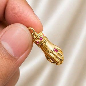 Pode incluir: Um pingente dourado em forma de mão, com um punho decorativo cravejado de pequenas pedras vermelhas redondas e uma única pedra vermelha retangular no dorso da mão. O pingente inclui um pequeno elo no topo para prender a uma corrente.