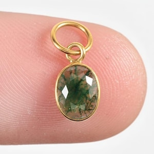 Puede incluir: Colgante dorado con una gema verde ovalada y facetada. La gema presenta un patrón similar al musgo. El colgante está suspendido de un pequeño lazo.
