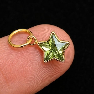 May include: A small star-shaped pendant featuring a faceted light green gemstone set in a polished gold-tone metal frame. The charm includes a circular gold-tone jump ring attached to the top point of the star, designed for easy attachment to chains.