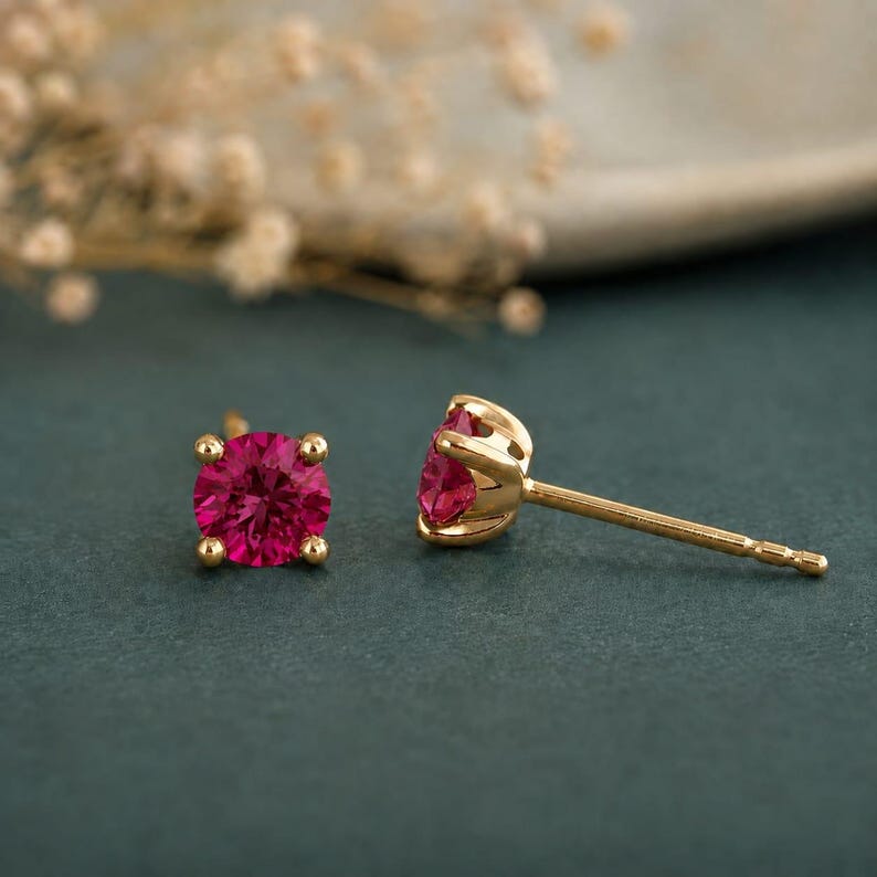 May include: A pair of stud earrings featuring round, vibrant pink gemstones set in polished yellow gold. One earring is shown from the front, displaying the circular stone and four-prong setting, while the other is positioned to show the side profile.