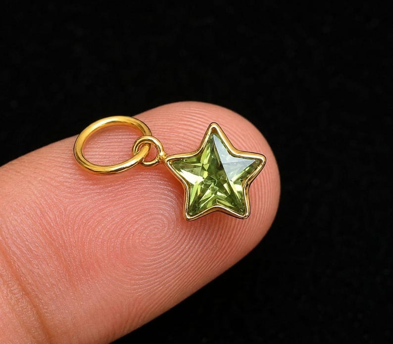 May include: A small star-shaped pendant featuring a faceted light green gemstone set in a polished gold-tone metal frame. The charm includes a circular gold-tone jump ring attached to the top point of the star, designed for easy attachment to chains.
