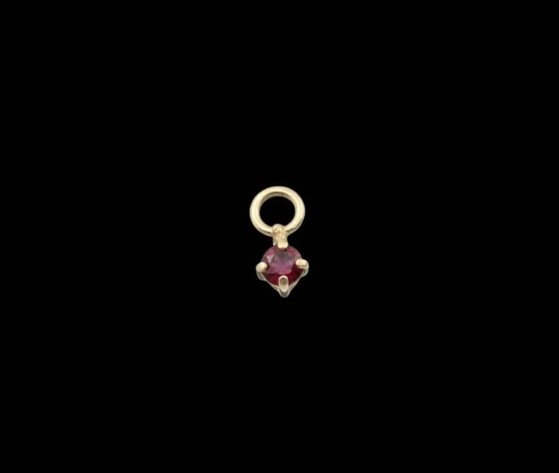 May include: A small, gold-coloured pendant with a round loop at the top and a red gemstone at the bottom. The gemstone is a deep, rich red colour and is set in a four-prong setting. The pendant is on a black background.