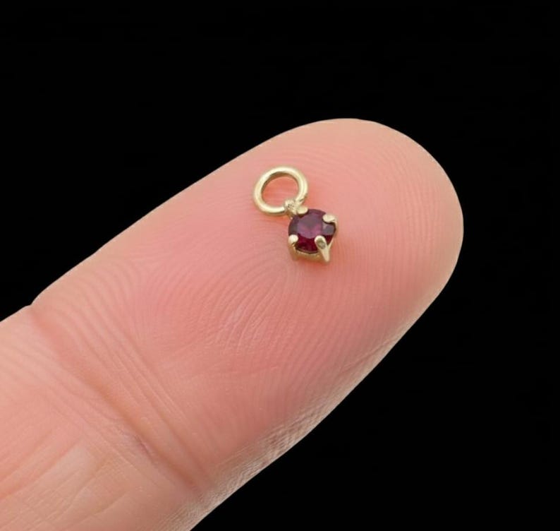 May include: A small, gold-coloured charm featuring a round, deep red gemstone. The charm has a loop at the top for attaching to a necklace or bracelet. The gemstone is set in a gold bezel.