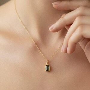 May include: A gold-colored necklace with a small, rectangular, dark green gemstone pendant. The pendant is suspended from a delicate chain. The necklace is worn on a person's neck.