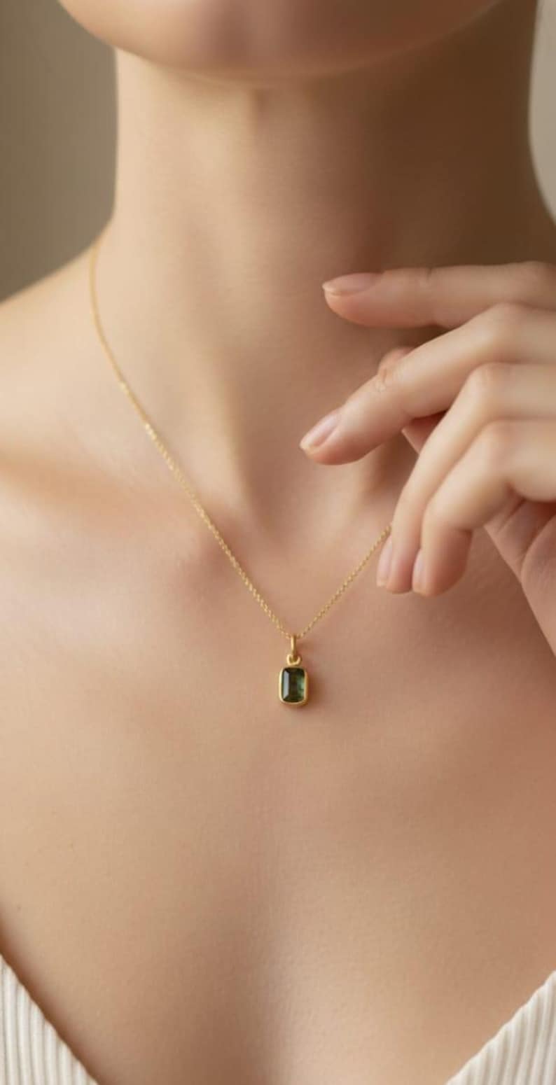 May include: A gold-colored necklace with a small, rectangular, dark green gemstone pendant. The pendant is suspended from a delicate chain. The necklace is worn on a person's neck.