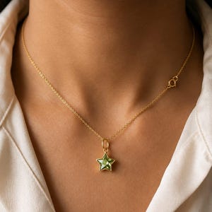 May include: A delicate gold chain necklace featuring a single star-shaped pendant with a faceted light green gemstone center. The necklace includes a thin gold link chain and a small circular spring ring clasp visible on the side of the neck.
