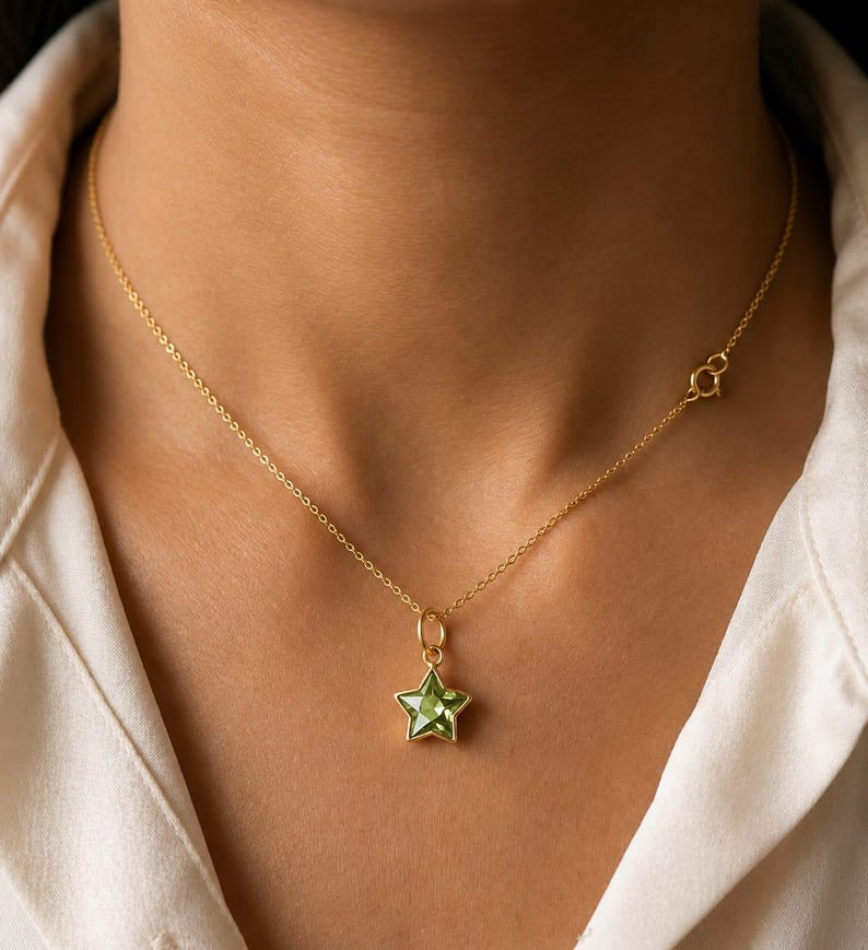 May include: A delicate gold chain necklace featuring a single star-shaped pendant with a faceted light green gemstone center. The necklace includes a thin gold link chain and a small circular spring ring clasp visible on the side of the neck.