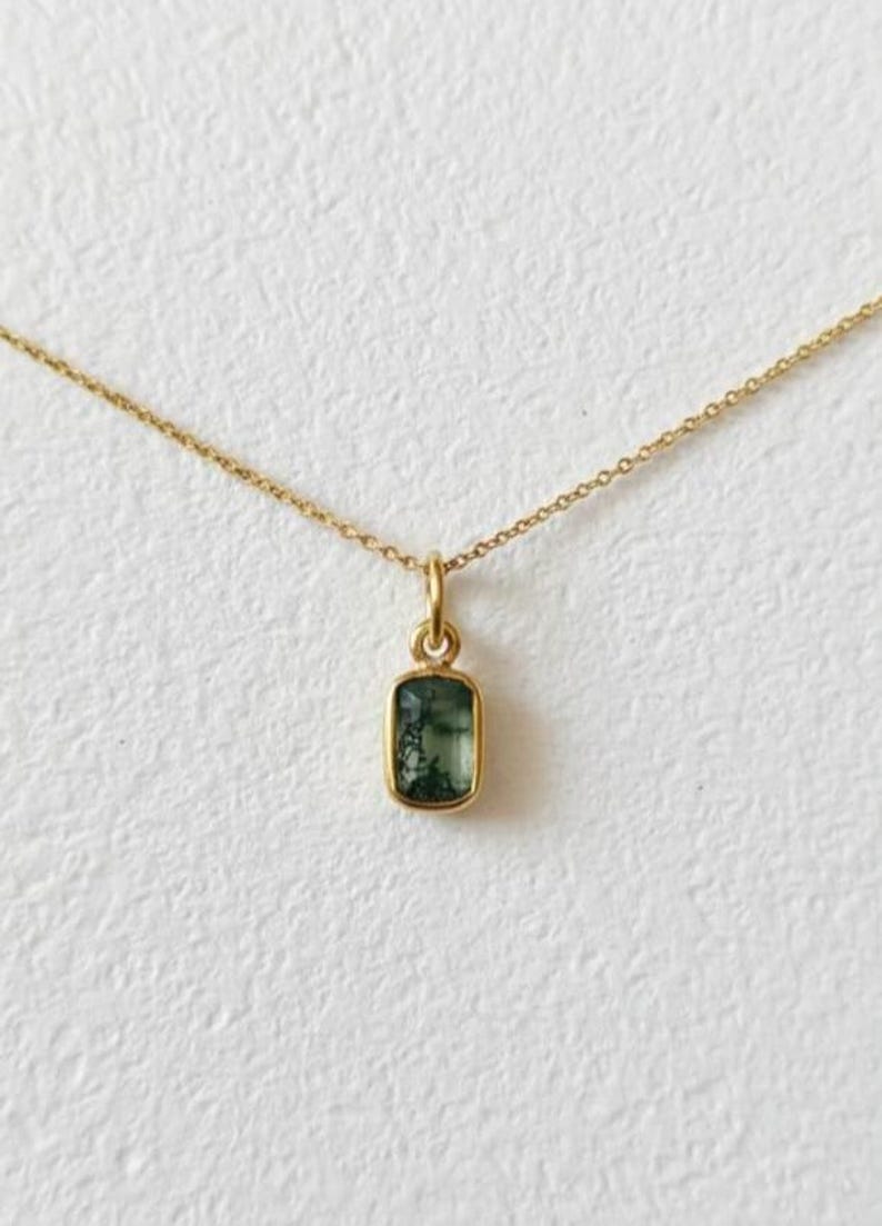 May include: A delicate gold necklace featuring a rectangular pendant with a green, moss-like design. The pendant is encased in a gold setting and hangs from a fine gold chain, set against a white background.