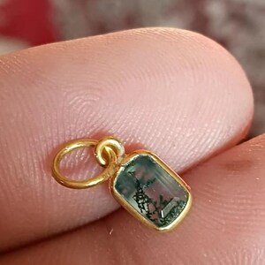 May include: A gold pendant with a rectangular, moss-green gemstone. The gemstone is set in a gold bezel and has a unique, natural pattern. The pendant is attached to a small gold loop.