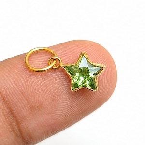 May include: A small star-shaped pendant featuring a faceted lime green gemstone set in a gold-toned metal frame. The charm includes a circular gold-toned jump ring attached to the top point of the star, designed for use as a jewelry component.