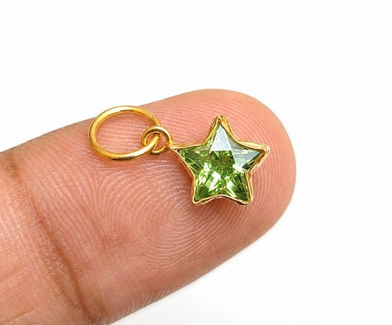 May include: A small star-shaped pendant featuring a faceted lime green gemstone set in a gold-toned metal frame. The charm includes a circular gold-toned jump ring attached to the top point of the star, designed for use as a jewelry component.