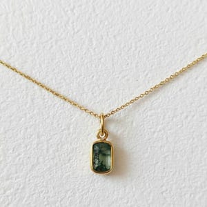 May include: A delicate gold necklace featuring a rectangular pendant with a green gemstone. The pendant is set in a gold bezel and hangs from a fine gold chain. The gemstone has a unique, natural pattern.