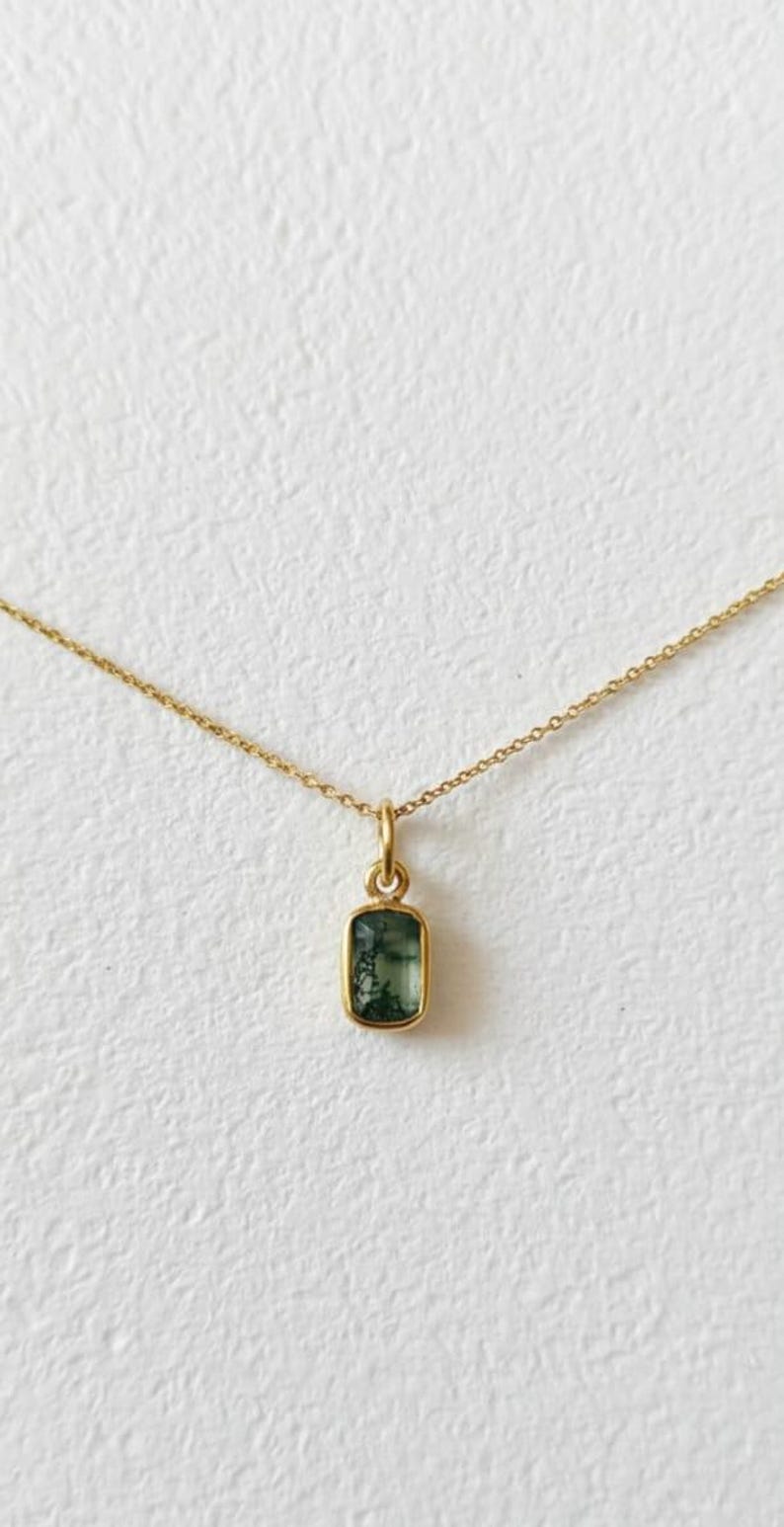 May include: A delicate gold necklace featuring a rectangular pendant with a green gemstone. The pendant is set in a gold bezel and hangs from a fine gold chain. The gemstone has a unique, natural pattern.