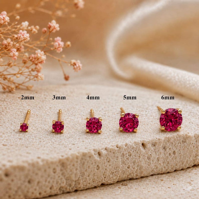 May include: Five gold stud earrings with round, vibrant pink stones, arranged in a row from smallest to largest. The sizes are labeled above each earring as 2mm, 3mm, 4mm, 5mm, and 6mm, indicating the diameter of the circular gemstone settings.