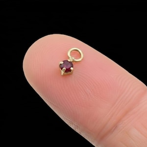 May include: A small, gold-coloured charm featuring a round, deep red gemstone. The charm is suspended from a small loop, and the gemstone is held in place by four prongs. The charm is shown against a blurred background.