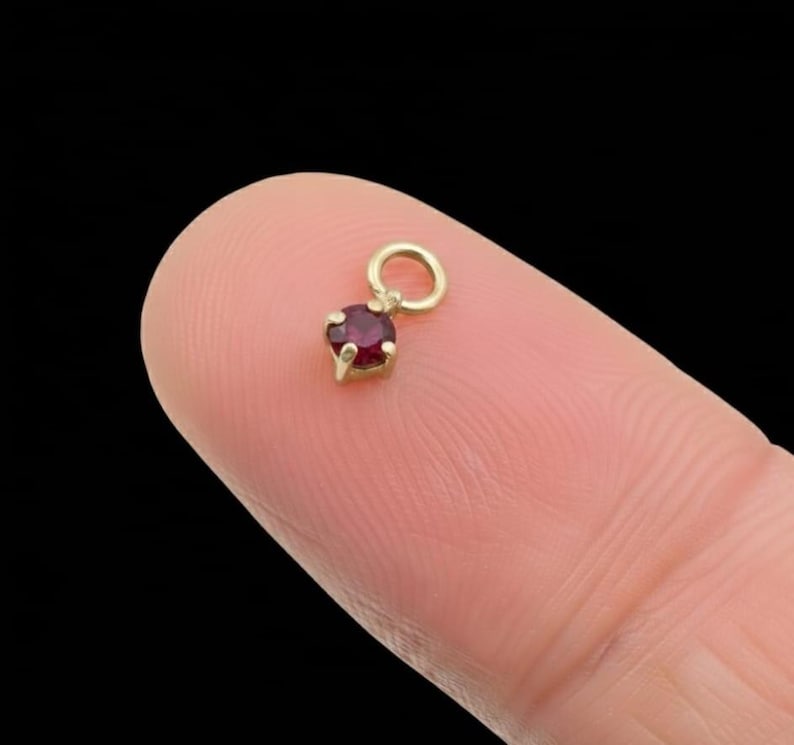 May include: A small, gold-coloured charm featuring a round, deep red gemstone. The charm is suspended from a small loop, and the gemstone is held in place by four prongs. The charm is shown against a blurred background.