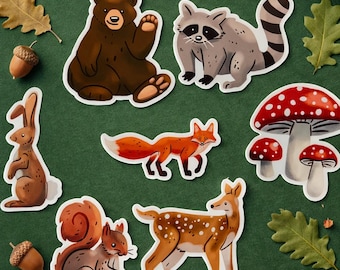 May include: A collection of woodland animal stickers, including a bear, raccoon, fox, squirrel, deer, and rabbit. The stickers are surrounded by acorns, oak leaves, a pine cone, and mushrooms on a green background.