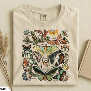 Może przedstawiać: Beżowa koszulka z kolorową grafiką motyli przedstawiającą różne gatunki. Projekt zawiera motyle w odcieniach niebieskiego, pomarańczowego, zielonego i czarnego. Koszulka ma okrągły dekolt i krótkie rękawy. Widoczna jest nazwa marki "Comfort Colors".