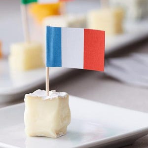 May include: A square of soft, creamy cheese is topped with a toothpick flag of the French tricolor: blue, white, and red. The cheese sits on a white plate, with other cheese cubes and flags in the background, suggesting a party or appetizer setting.