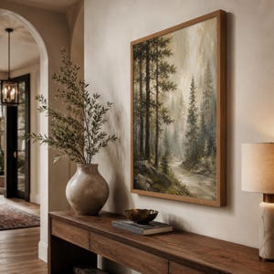 May include: A rectangular framed wall art print depicting a forest landscape with tall pine trees, a winding river, and rocky terrain in muted earth tones of green, brown, and beige, displayed in a wooden frame on a neutral-colored wall.