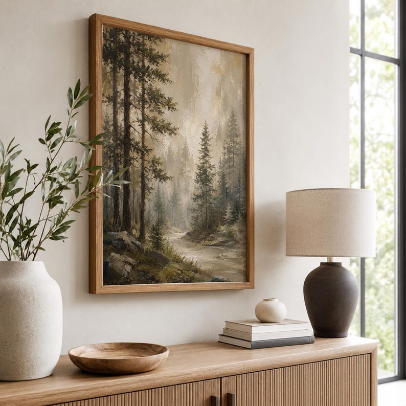 May include: A framed wall art print depicting a misty forest landscape with tall pine trees and a winding stream in muted earth tones of brown, green, and beige, displayed in a wooden frame with a natural finish and visible wood grain texture.