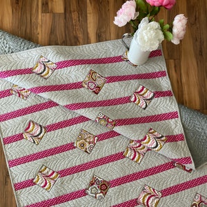 May include: A gray and pink quilt with a striped pattern and diamond-shaped fabric squares with floral designs. The quilt is quilted with a curved pattern.