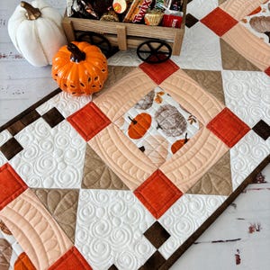 May include: A fall-themed table runner with a quilted design featuring pumpkins and autumn leaves. The runner is paired with a decorative pumpkin, a white pumpkin, and a wooden wagon filled with candy.