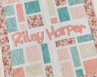 May include: A pink and white baby quilt with a floral print border. The quilt is made up of squares of different fabrics, including a light blue fabric with a swirl pattern, a pink fabric with a floral pattern, and a white fabric with a gold dot pattern. The quilt is personalized with the name "Riley Harper" in a pink font.