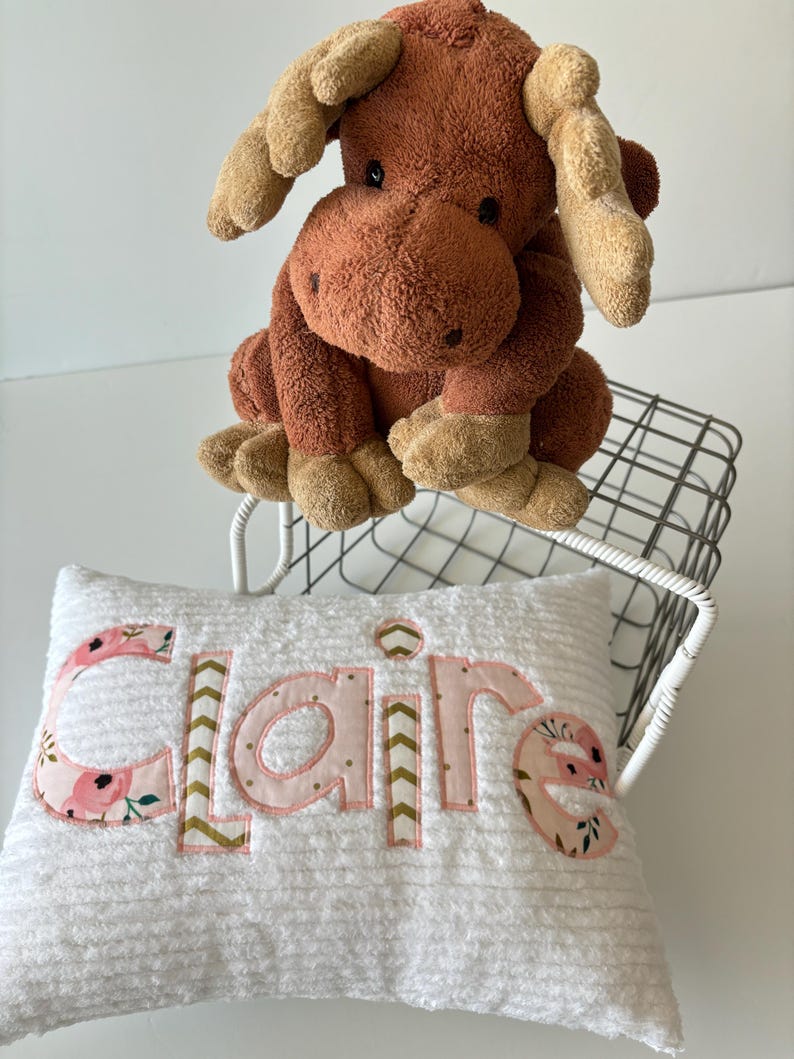 May include: A white pillow with the name "Claire" embroidered in pink letters with floral and chevron patterns. A brown plush moose toy sits on a metal wire basket.