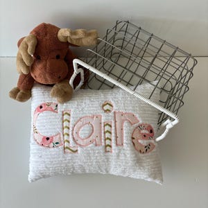 May include: A white pillow with the name "Claire" embroidered in pink and gold letters with floral accents. The pillow is sitting on a wire basket with a brown plush moose toy.