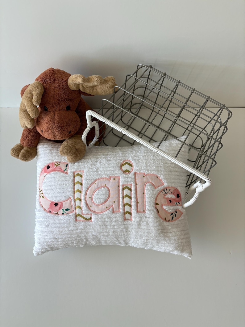 May include: A white pillow with the name "Claire" embroidered in pink and gold letters with floral accents. The pillow is sitting on a wire basket with a brown plush moose toy.