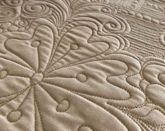 May include: A beige quilted table runner with a decorative floral pattern. The pattern is made up of swirls and circles.