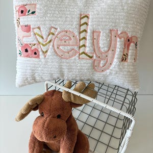 May include: A white plush pillow with the name "Evelyn" spelled out in pink fabric letters with gold accents. The letters are decorated with a floral pattern. A brown stuffed moose sits in front of the pillow.