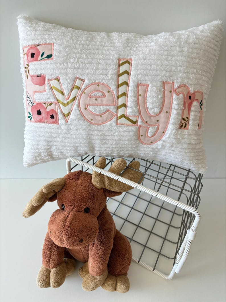 May include: A white plush pillow with the name "Evelyn" spelled out in pink fabric letters with gold accents. The letters are decorated with a floral pattern. A brown stuffed moose sits in front of the pillow.