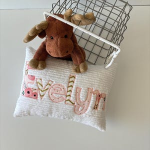 May include: A white plush pillow with the name "Evelyn" embroidered in pink letters with floral accents. A brown moose stuffed animal is peeking out from behind the pillow.