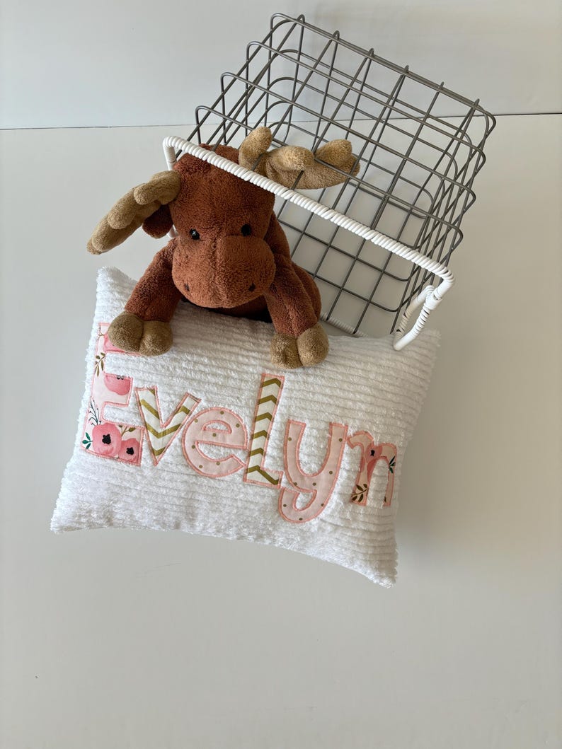 May include: A white plush pillow with the name "Evelyn" embroidered in pink letters with floral accents. A brown moose stuffed animal is peeking out from behind the pillow.