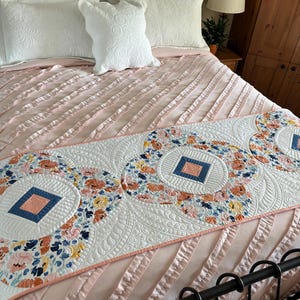 May include: A white and pink bed with a white quilted bed runner featuring a floral pattern and blue and orange accents. The bed runner has a scalloped edge and is placed on a pink bedspread with ruffles.