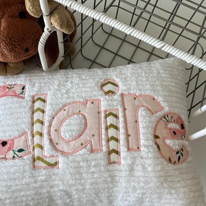May include: A white pillow with the name "Claire" embroidered in pink and gold fabric. The letters are outlined in pink and the fabric features a floral pattern and a chevron pattern.