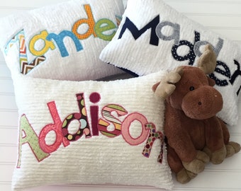May include: Three white pillows with the names "Tanden", "Mader", and "Addison" spelled out in colorful fabric letters. A brown moose stuffed animal is lying on top of the "Addison" pillow.