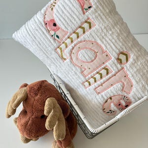 May include: White plush pillow with the name "Claire" embroidered in pink letters with a floral and chevron pattern. The pillow is in a wire basket with a brown plush moose toy.